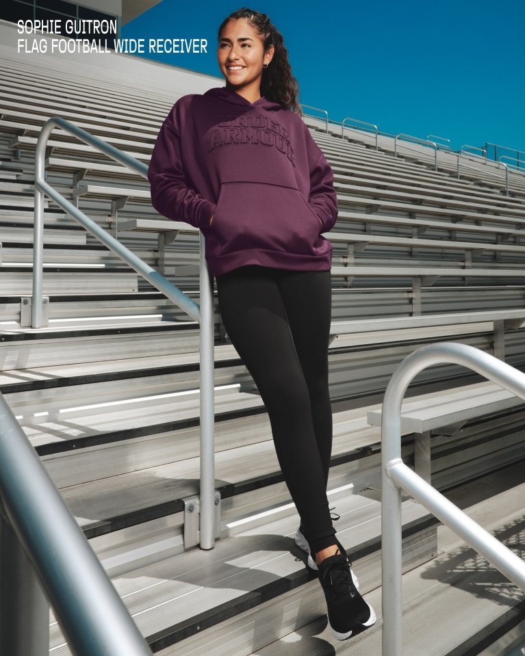 Athlete in Under Armour hoodie and leggings stands on stadium bleachers with hands in pocket.