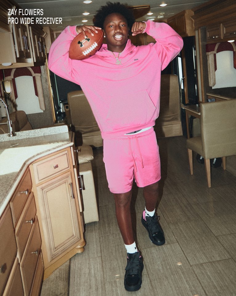 Athlete in pink Under Armour hoodie and shorts holds a football on their shoulder in a kitchen.