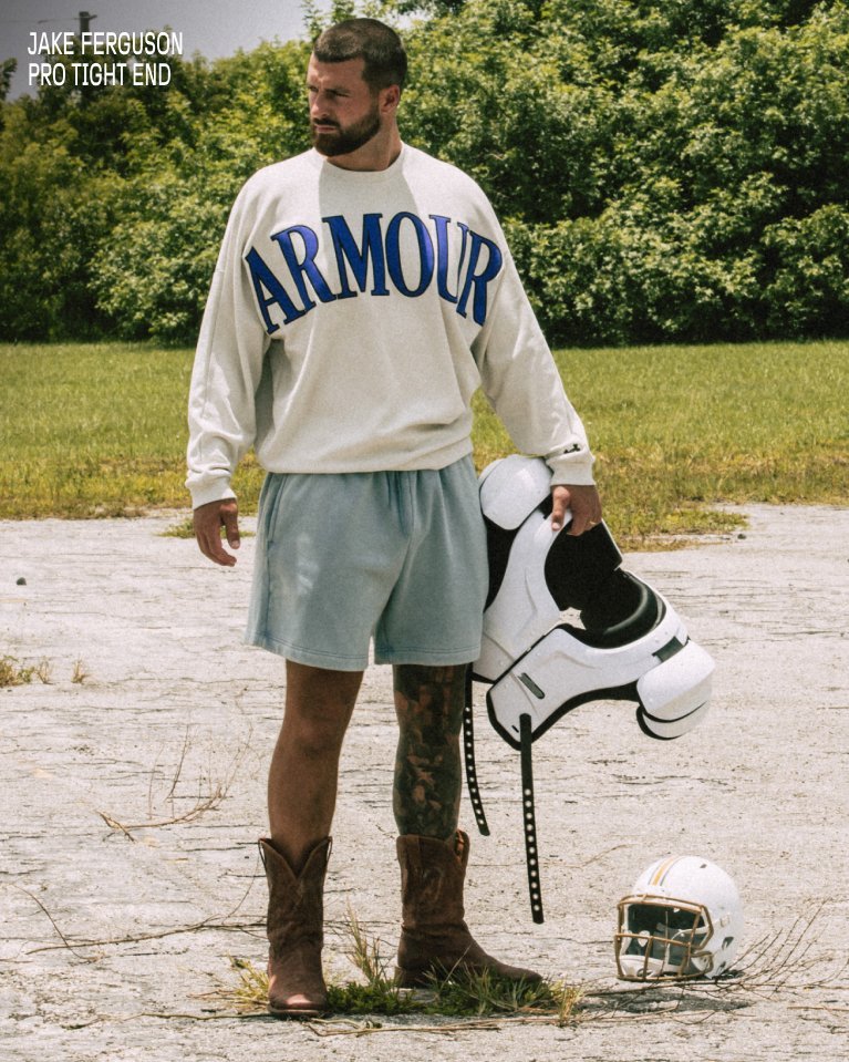 Athlete stands on a field wearing an Under Armour sweatshirt and holds football pads with a helmet on the ground.