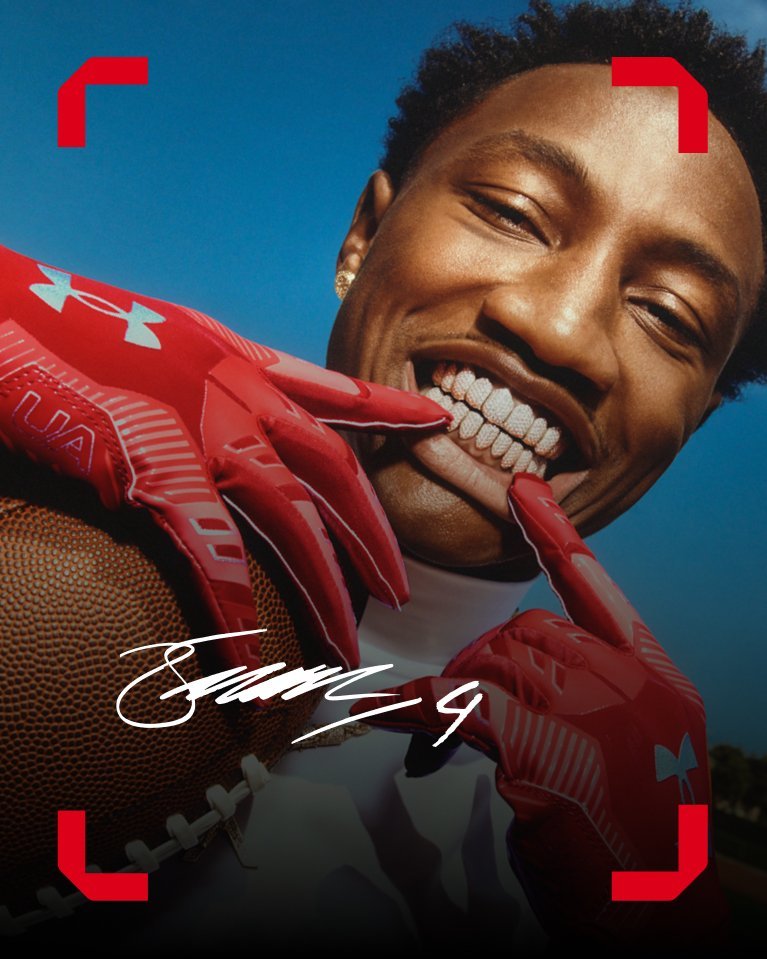 Athlete smiles while holding a football and wearing red Under Armour gloves.