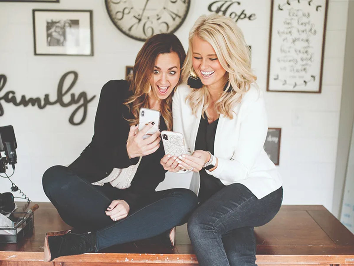 Two friends laughing together over a phone on a sunny patio.