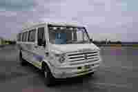 A white Mahindra Force van with "TEJAS" on the windshield, parked on a concrete road under a cloudy sky.