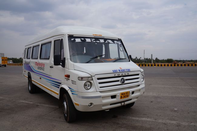 A white Mahindra Force van with "TEJAS" on the windshield, parked on a concrete road under a cloudy sky.