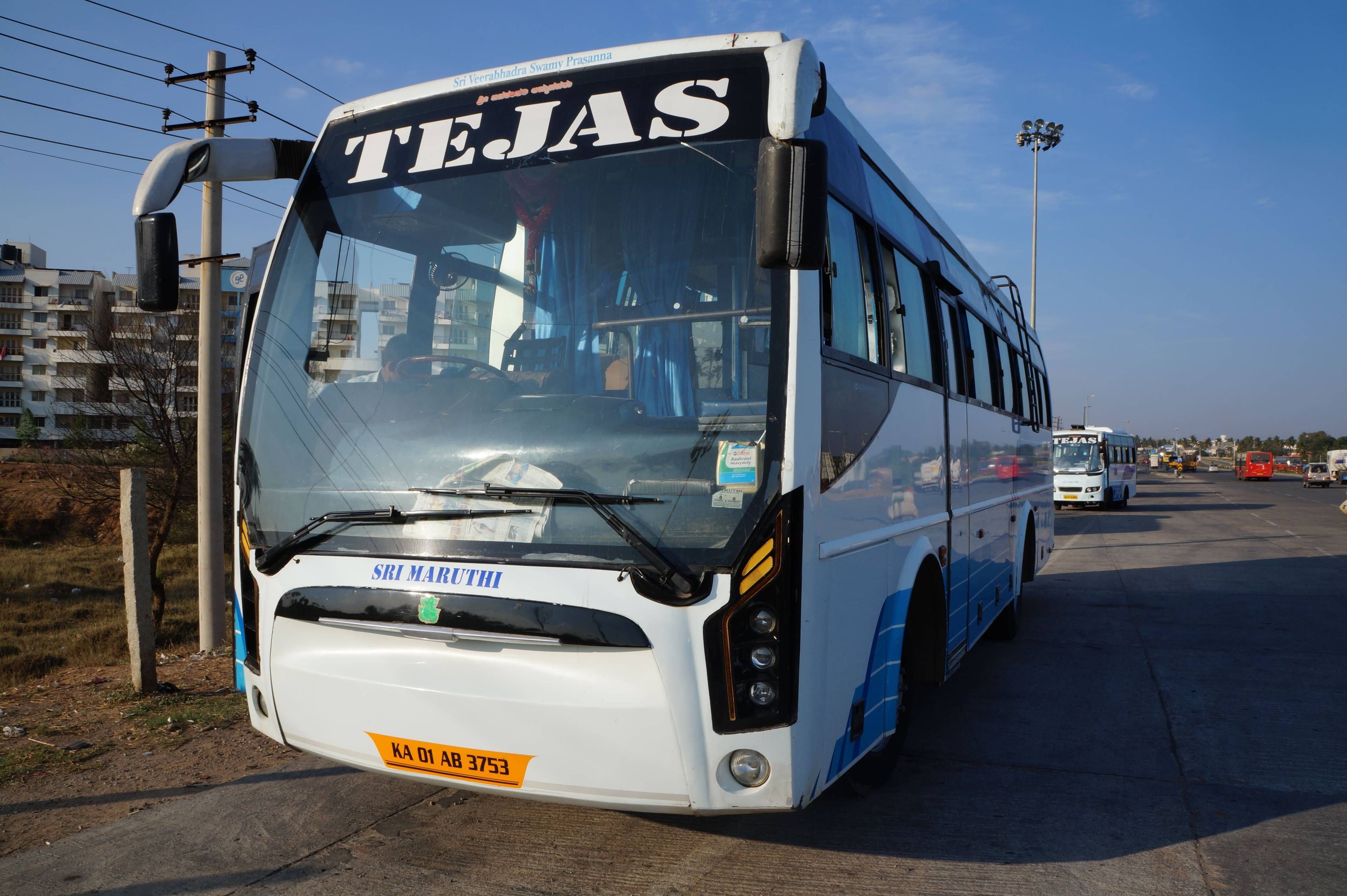 A white and blue bus labeled "TEJAS" parked on a roadway near tall poles, with buildings visible in the background.