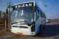 A white bus labeled "TEJAS" parked at a bus station, with a blue sky and nearby buildings in the background.