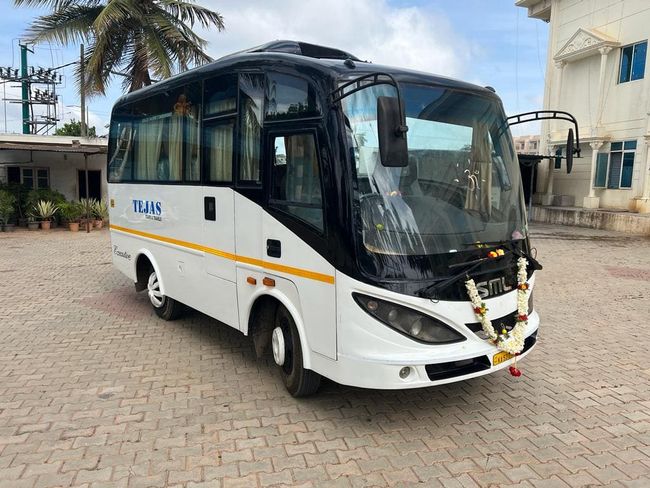 A white and black SML bus adorned with floral garlands parked on a paved area, surrounded by palm trees and blue sky.