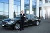 A confident woman in a black suit stands with arms crossed beside a sleek black luxury car in front of a modern building.