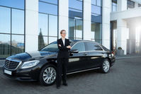 A confident woman in a black suit stands with arms crossed beside a sleek black luxury car in front of a modern building.