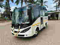 A white minibus with a floral garland parked on a tiled surface, surrounded by palm trees and a building in the background.