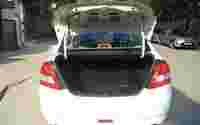 A white car with its trunk open, revealing a spacious empty trunk area on a street with parked vehicles in the background.