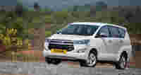 A white Toyota MPV parked on a gravel road with a green landscape and mountains in the background.