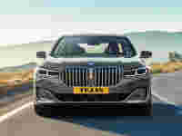 A sleek gray BMW 7 Series car drives on a winding road, with mountains in the background and a clear sky above.
