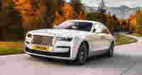 Luxurious white car driving on a winding road surrounded by colorful autumn trees and mountains in the background.