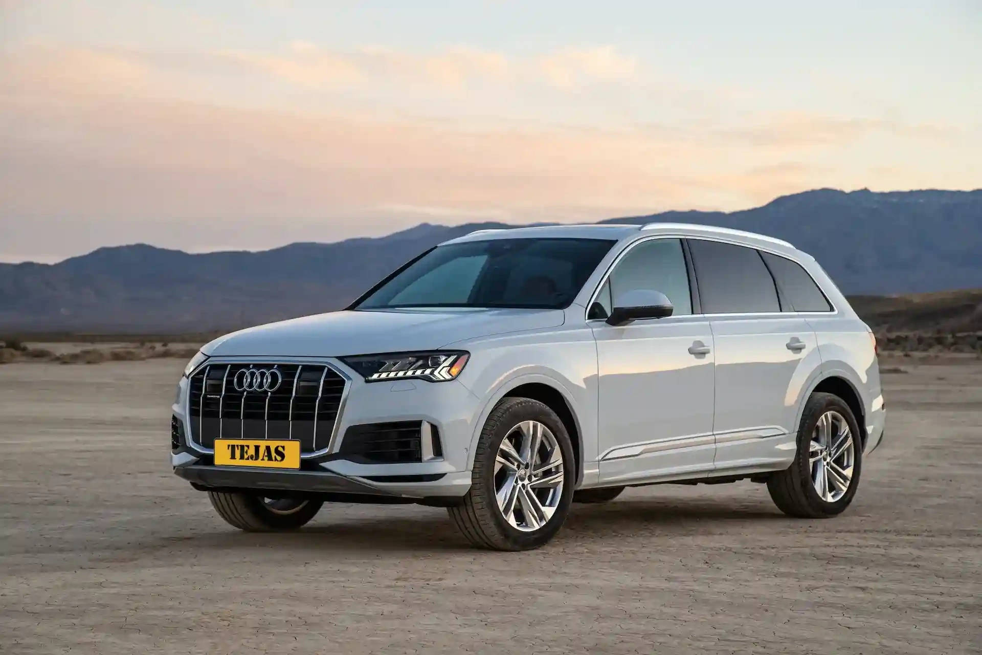 Silver Audi Q7 SUV parked on dry desert land with mountains in the background and a colorful sunset sky.
