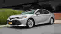 A silver Toyota Camry parked on a pavement beside tall green grass and a modern building.