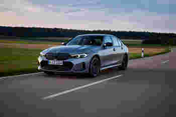 A silver BMW sedan speeds along a scenic rural road surrounded by green fields and trees under a cloudy sky.