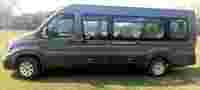 A dark gray van parked on green grass, featuring multiple seating rows and large windows, with a city skyline visible in the background.