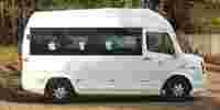 White van parked on a dirt area surrounded by trees, featuring large windows and visible seating inside.