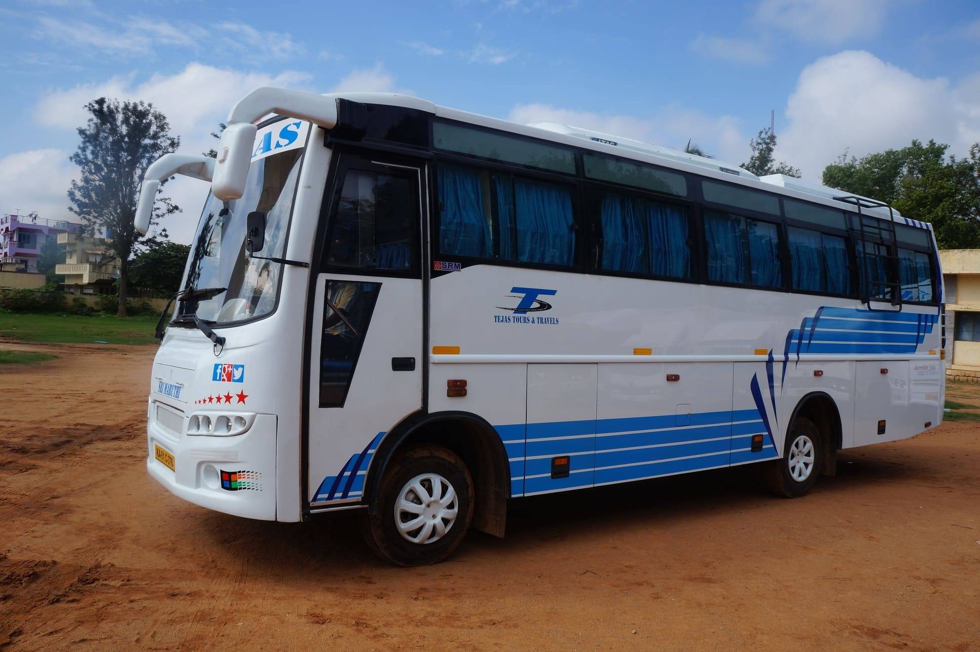 A white and blue tour bus parked on a dirt road, with trees and buildings in the background under a partially cloudy sky.