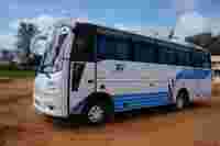 A white and blue tour bus parked on a dirt road, with trees and buildings in the background under a partially cloudy sky.