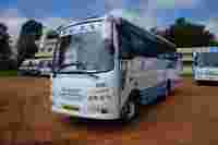 A white bus with blue accents, labeled "TEJAS," parked on a dirt area near buildings and other buses.