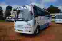 A white bus with blue accents, labeled "TEJAS," parked on a dirt area near buildings and other buses.
