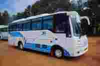 A white and blue tour bus parked on a dirt lot, surrounded by greenery and showing a driver inside.