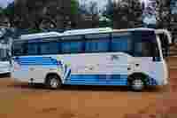 A white travel bus with blue accents parked on a dirt surface, surrounded by trees and another bus in the background.