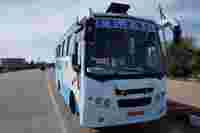 A blue and white bus named "Tejas" parked beside a roadside with buildings and trees in the background under a clear sky.