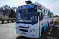 A white and blue bus labeled "TEJAS" parked along a road near greenery and a commercial building, under a clear sky.