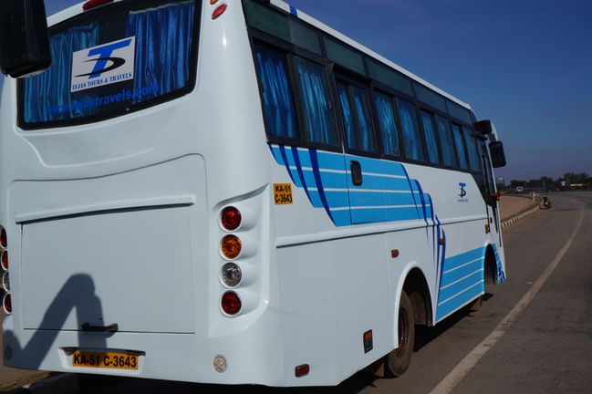 A white and blue bus labeled "TEJAS" parked along a road near greenery and a commercial building, under a clear sky.