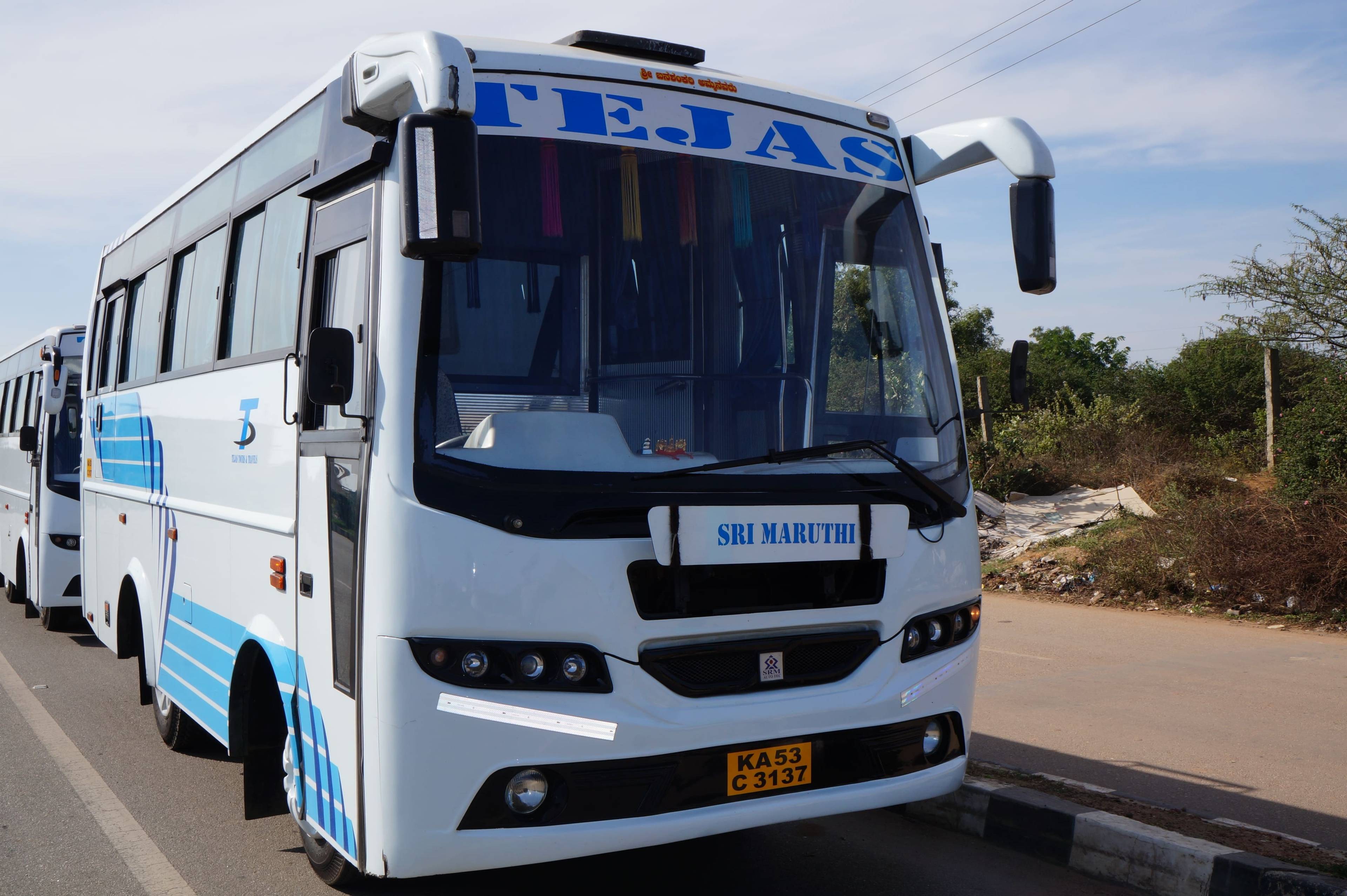 White bus with blue accents labeled "TEJAS" parked along a road, surrounded by vegetation and a clear sky.