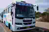 A blue and white bus labeled "TEJAS" parked beside a road, with trees and a patch of land in the background.