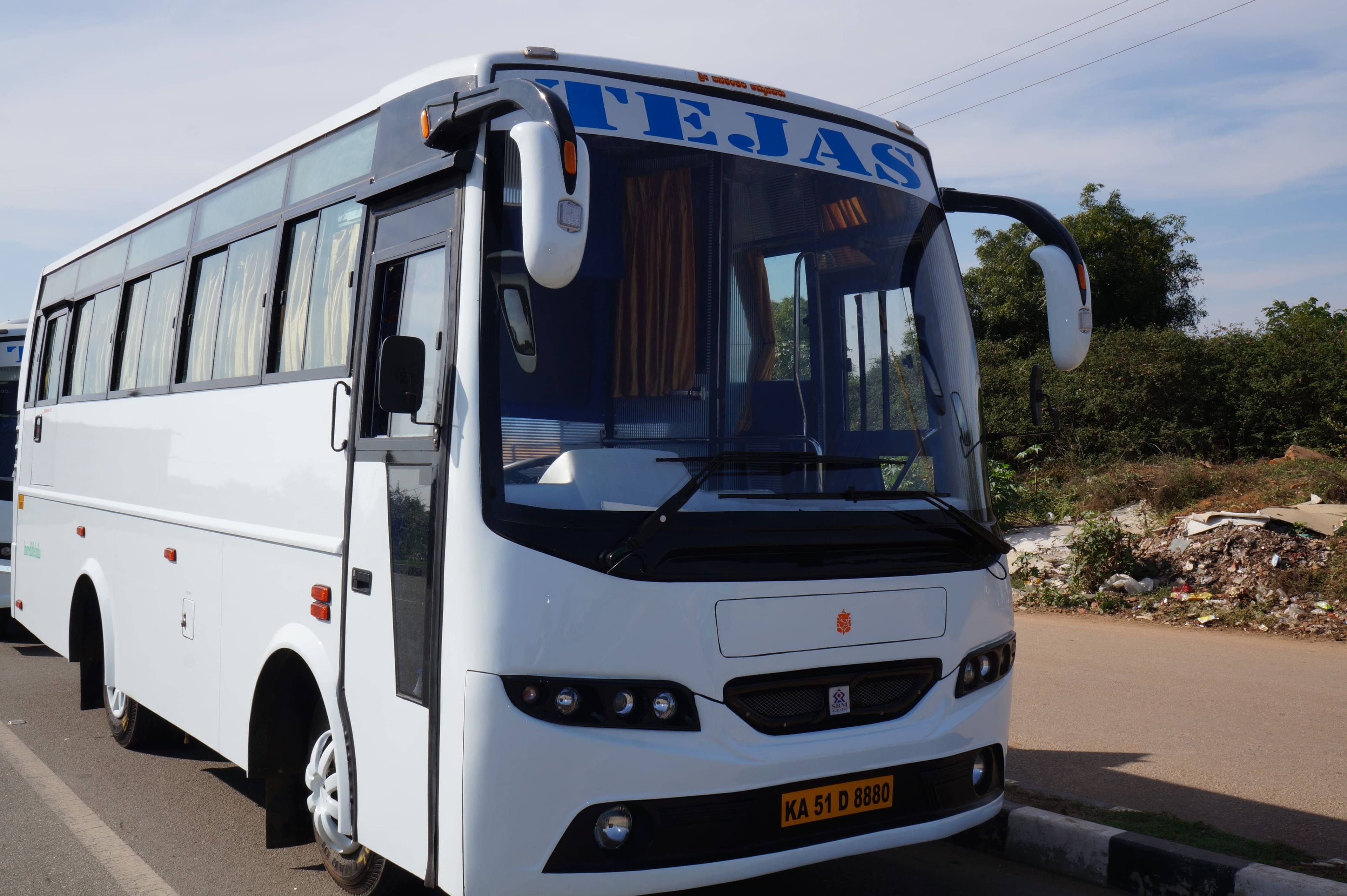 A modern white bus with the name "TEJAS" displayed prominently on the front, parked beside a road with greenery and debris nearby.