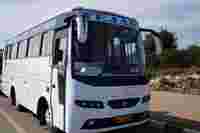 A white bus labeled "TEJAS" parked along a road, with a clear sky and green vegetation in the background.