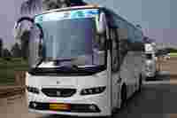 White passenger bus with a blue "T.JAS" logo parked on a wide road, palm trees and buildings visible in the background.