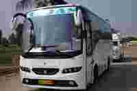 A white bus with "T.J.A.S" on the front, parked on a concrete surface surrounded by palm trees and other vehicles.