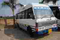 A white SML bus named "Tejas" parked near a palm tree, surrounded by a rural landscape.