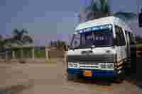 Bus labeled "TEJAS" parked on a concrete surface, surrounded by palm trees and open fields under a clear sky.