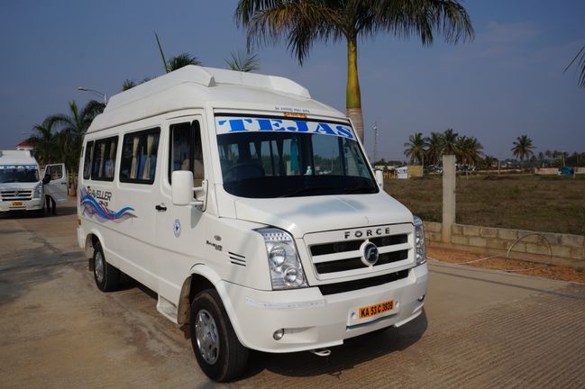 A white traveler van with "TEJAS" on the windshield is parked on a road, surrounded by palm trees and open land.