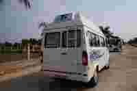 White travel van parked on a paved road, surrounded by palm trees and green landscaping, with another vehicle in the background.