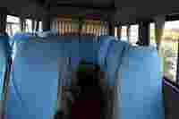 Empty bus interior with rows of seats covered in blue fabric, red carpet on the floor, and light streaming through windows.