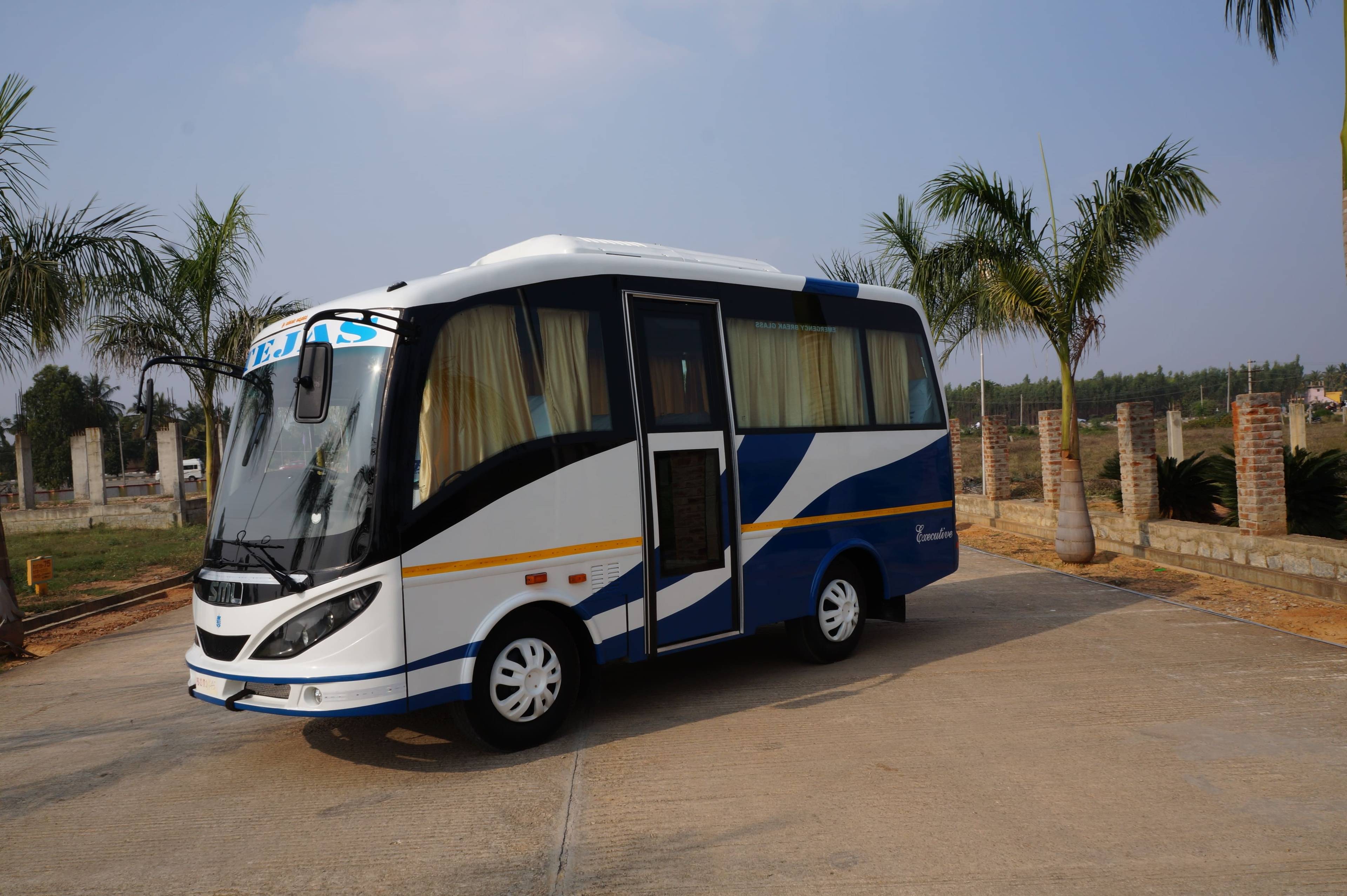 White and blue travel bus parked on a concrete road, surrounded by palm trees and an open landscape in the background.
