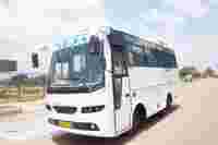 A white bus with the word "TEJA" on the front, parked on a road with green landscaping and blue skies in the background.