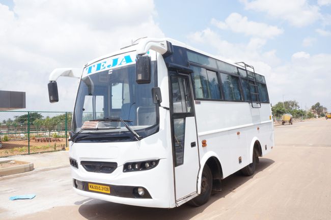 A white tour bus with large windows and curtains parked by the roadside, surrounded by greenery under a clear sky.