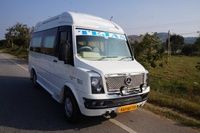 A white van with "TEJAS" on the windshield parked on a roadside, surrounded by greenery and distant hills.