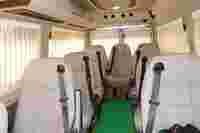 Interior of a van featuring multiple beige seats with seat belts, green carpet, and large windows with light-colored curtains.