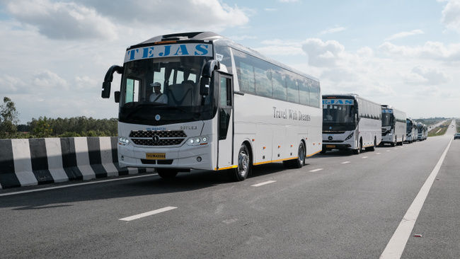 Two white luxury buses parked on a dirt lot, displaying "TEJAS" and "Travel With Dreams" branding. Greenery in background.