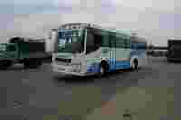 A blue and white bus labeled "TEJAS" drives past a green truck on a wide road under a cloudy sky.