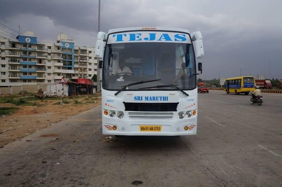 A white bus labeled "TEJAS" and "SRI MARUTHI" is parked on a road near residential buildings under a cloudy sky.
